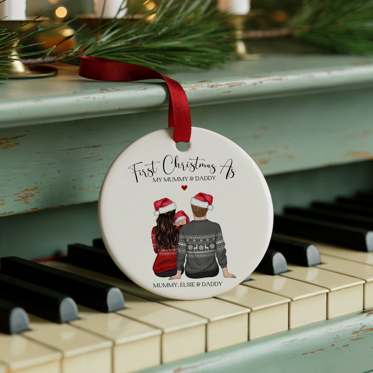 A round white ceramic Christmas ornament with a red ribbon, featuring a custom illustration of a couple in holiday sweaters, hanging on a distressed teal piano.
