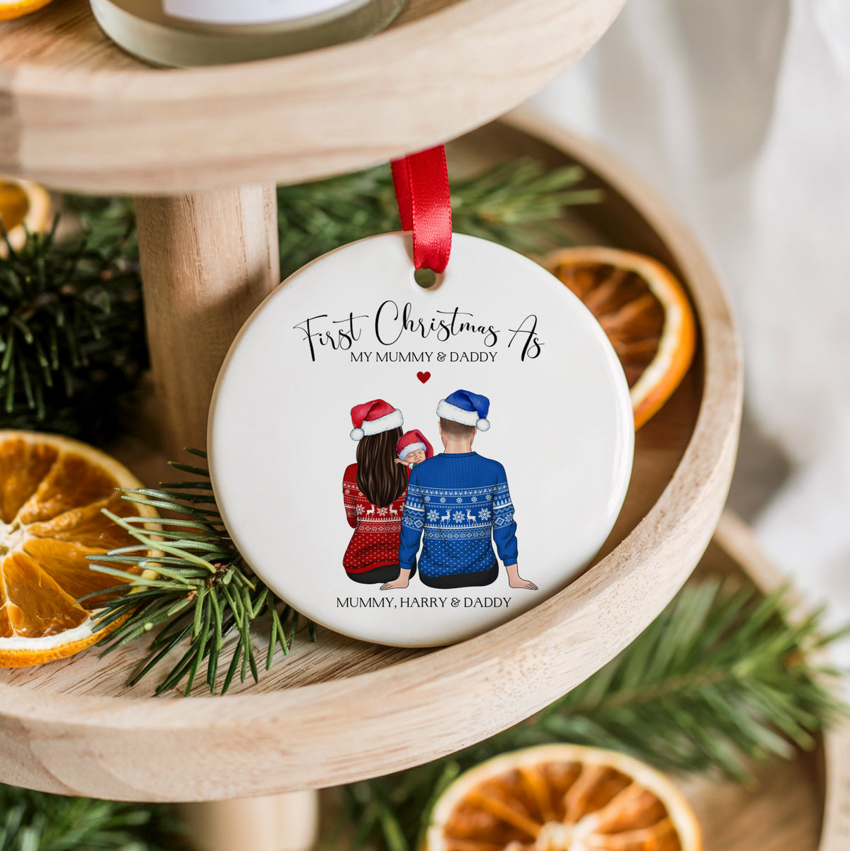 A round white ceramic Christmas ornament hanging from a red ribbon, printed with a personalized illustration of a couple seen from behind wearing matching sweaters, with the text 'First Christmas As MY MUMMY & DADDY' and 'MUMMY, HARRY & DADDY'. The ornament is styled on a wooden tiered stand with pine branches and dried orange slices.