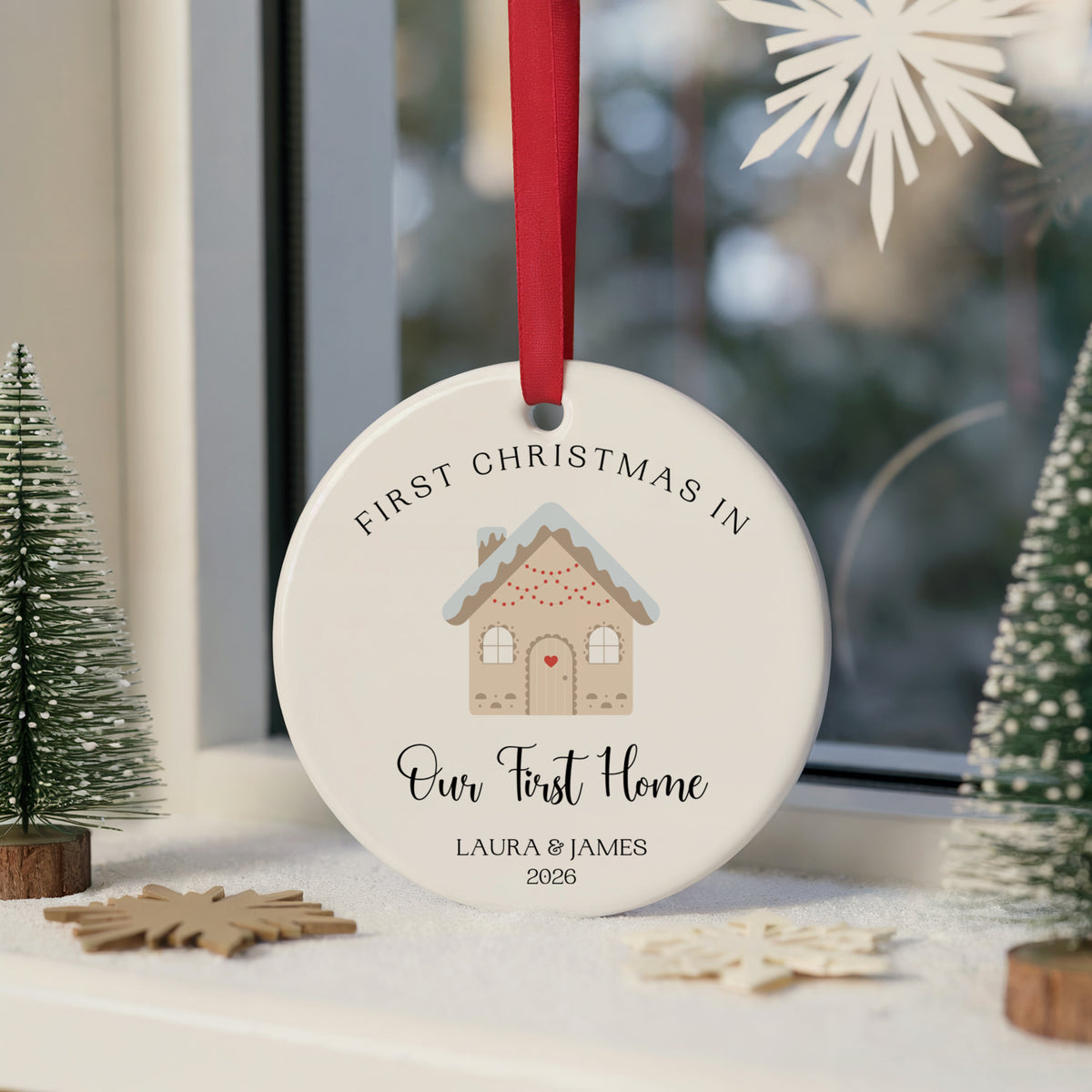 A personalized round white Christmas ornament hanging from a red ribbon, printed with a gingerbread house illustration and the text 'FIRST CHRISTMAS IN Our First Home LAURA & JAMES 2025'. The ornament is styled on a windowsill with miniature trees and wooden snowflake decorations.