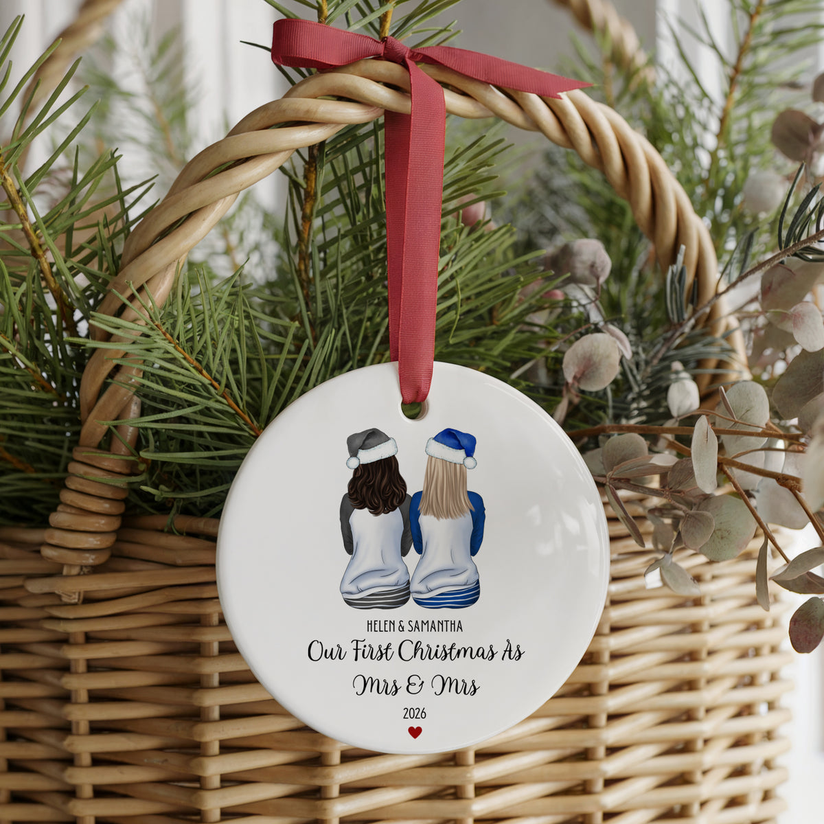 A round white ceramic Christmas ornament with a red ribbon hanging from a wicker basket, decorated with an illustration of two women seen from behind and the personalized text 'HELEN & SAMANTHA Our First Christmas As Mrs & Mrs 2026'.