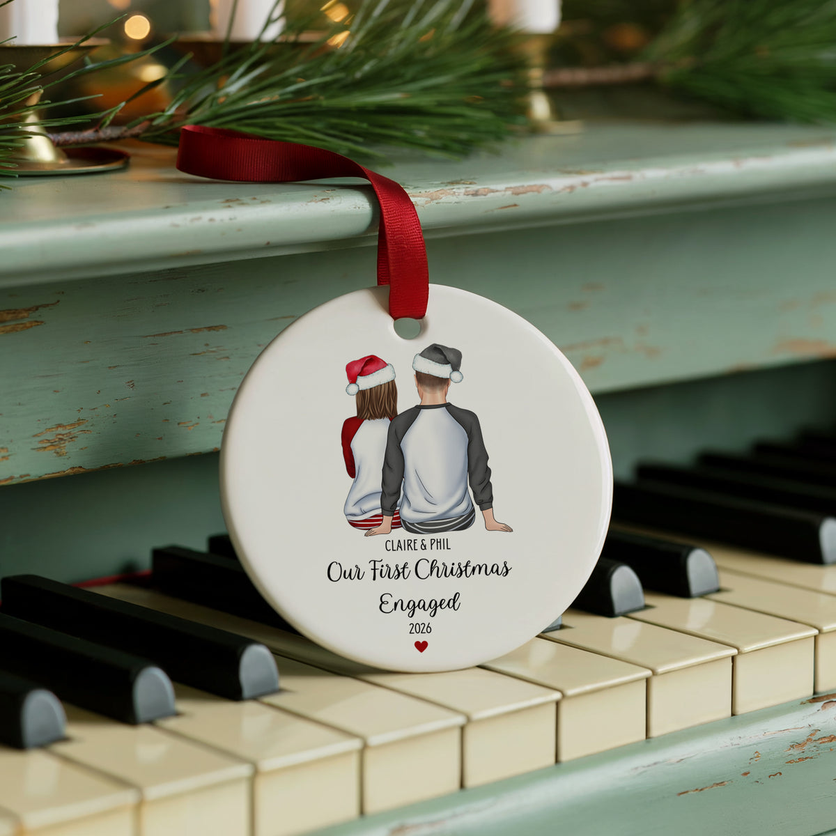 A round white Christmas ornament with a red ribbon, featuring an illustration of a couple from behind wearing Santa hats, hanging on a distressed teal piano. The ornament is personalized with the text 'CLARK & PEE Our First Christmas Engaged 2023'.