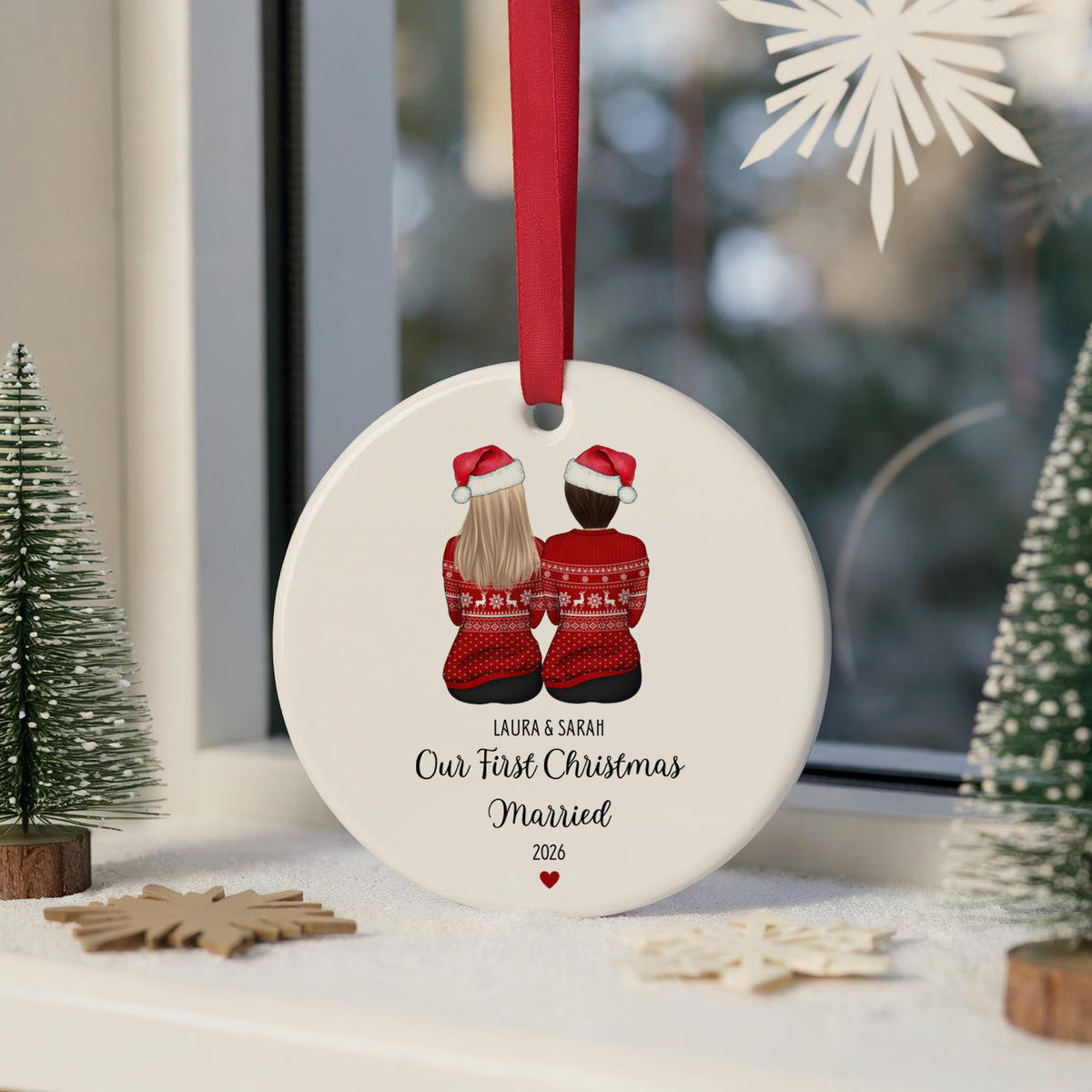 A personalized round white Christmas ornament hanging from a red ribbon, featuring an illustration of a couple from behind in red sweaters and Santa hats, with the text 'LAURA & SARAH Our First Christmas Married 2026'. The ornament is styled on a snowy windowsill with miniature trees and wooden star decorations.