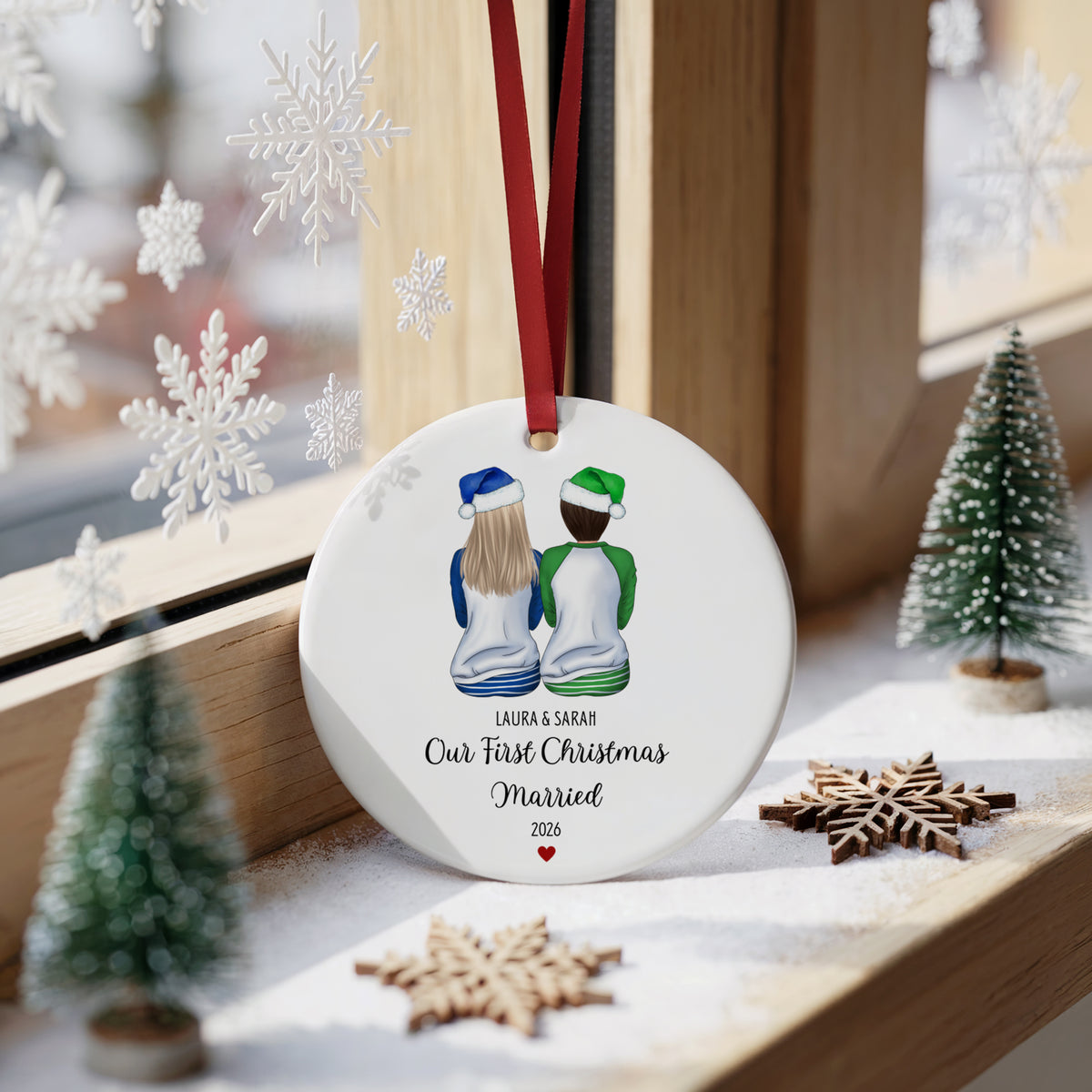 A round white Christmas ornament hanging on a wooden windowsill, printed with an illustration of a couple from behind wearing Santa hats and the text 'LAURA & SKRAM Our First Christmas Married 2026.' The ornament is decorated with a red hanging ribbon and is surrounded by holiday props including small trees, wooden snowflakes, and white snowflake decorations.