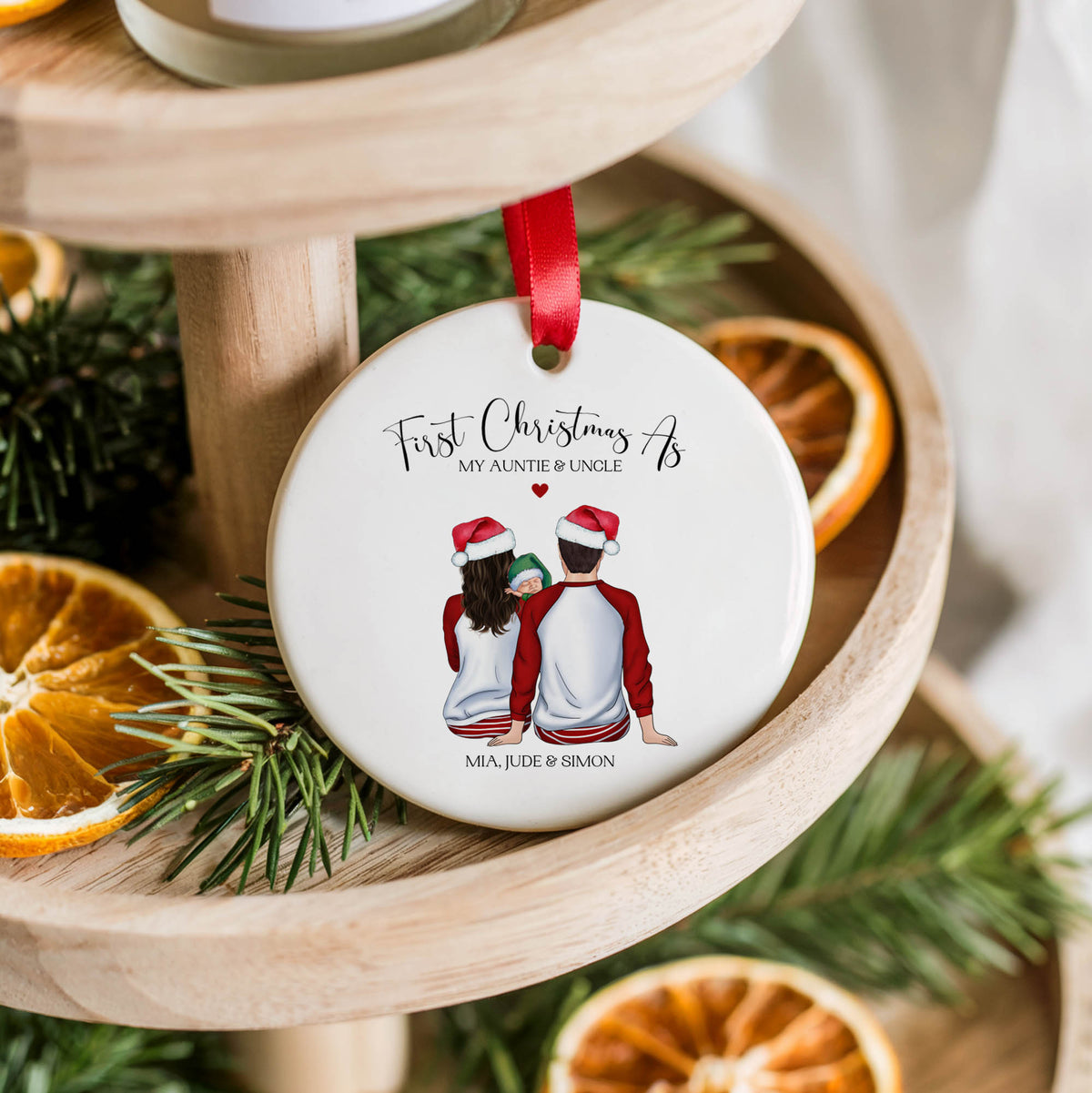 A personalized round ceramic Christmas ornament hanging on a wooden tiered stand, decorated with a red ribbon. The white ornament features a printed illustration of a couple wearing Santa hats and the text 'First Christmas My Nontie & Uncle' along with names at the bottom.