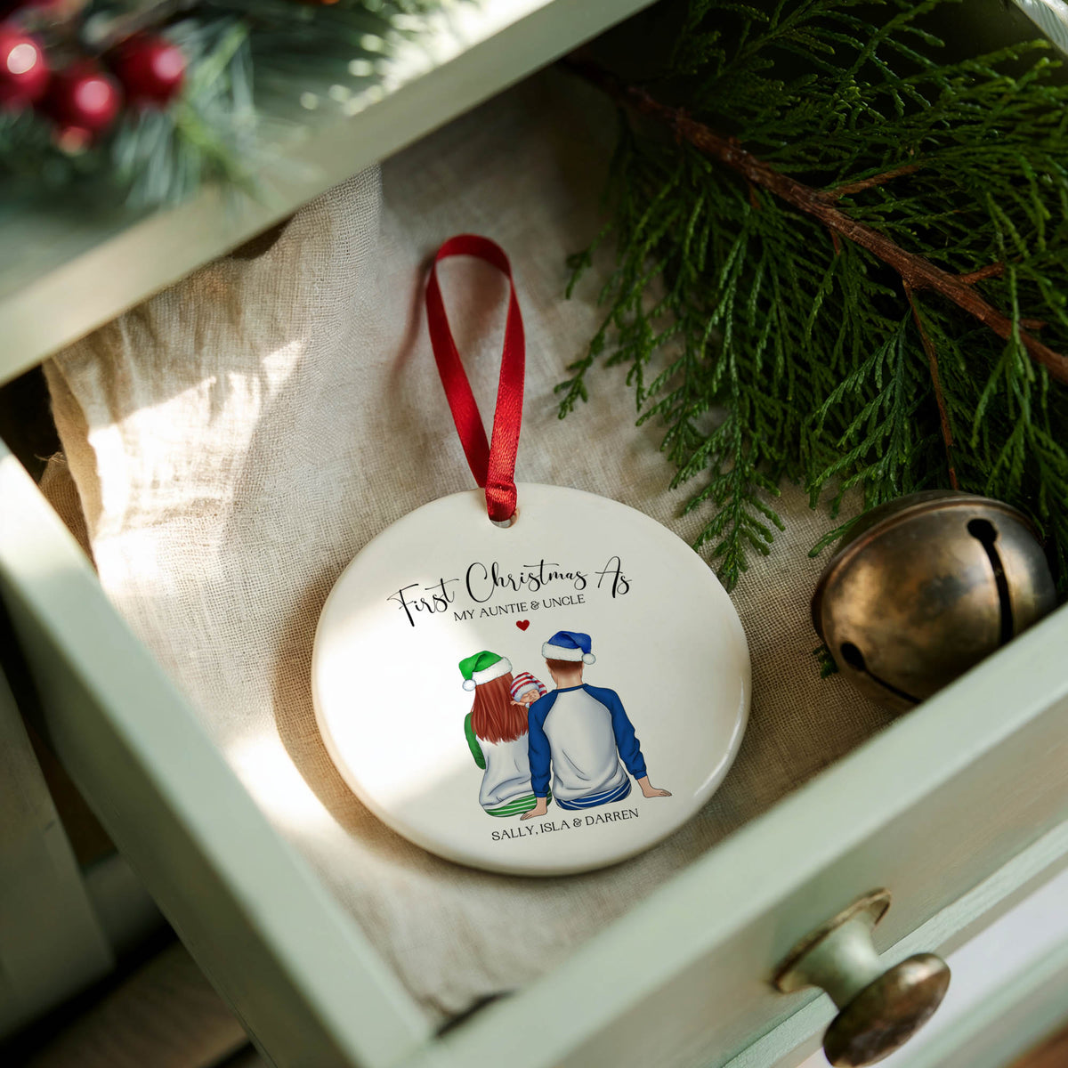 A round white ceramic Christmas ornament with a red hanging ribbon, featuring a custom illustration of a father and son in baseball uniforms and the text 'First Christmas As' and 'BULLSEYE GAMES'. The ornament is styled in a light green drawer lined with beige fabric, surrounded by pine branches and a small brass bell.