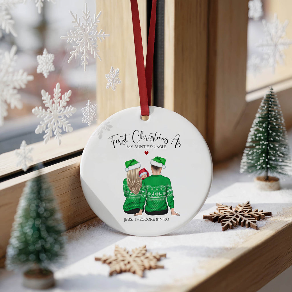 A personalized round Christmas ornament hanging on a wooden windowsill. The ornament is white with a red hanging ribbon and features an illustration of a mother and son wearing green sweaters, with the text 'First Christmas As MY AUNTIE OR UNCLE' and names printed below.