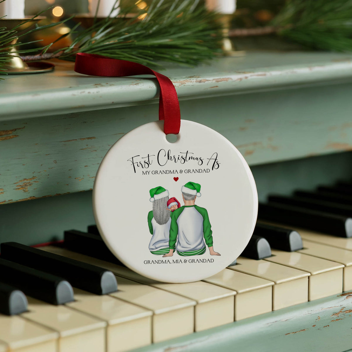 A personalized round ceramic Christmas ornament hanging from a red ribbon on a vintage teal piano. The ornament features an illustration of a grandfather and grandmother wearing green and white holiday sweaters, with the text 'First Christmas As MY GRANDMA & GRANDPAD' and 'GRANDMA, MIA & GRANDPAD'.