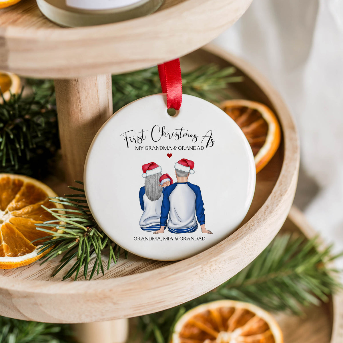 A round white ceramic Christmas ornament hanging from a red ribbon, printed with the text 'First Christmas As MY GRANDMA & GRANDPAD' and an illustration of a couple and their grandson seen from behind. The ornament is styled on a wooden tiered stand with pine branches and dried orange slices.