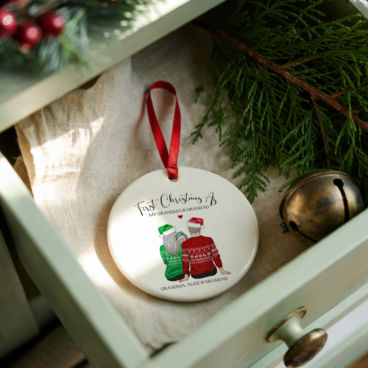 A round white Christmas ornament with a red ribbon loop, featuring a printed illustration of a grandfather and grandmother in holiday sweaters and the text 'First Christmas With My Grandpa & Grandma', displayed in an open drawer with pine branches, a bell, and a beige cloth.