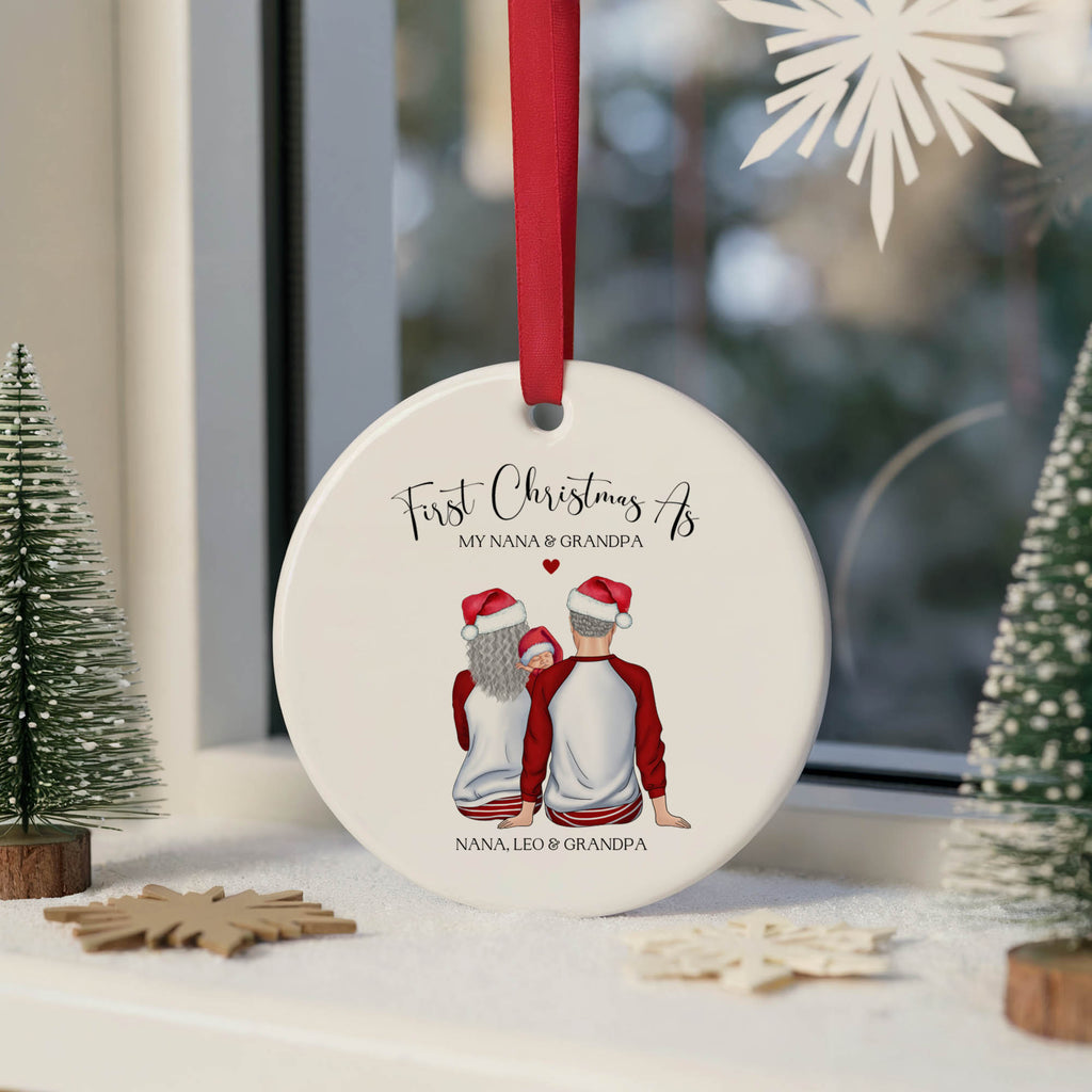 A personalized round white ceramic Christmas ornament hanging from a red ribbon, featuring an illustration of a grandfather and his two grandchildren sitting together with the text 'First Christmas As MY NANA & GRANDPA' and 'NANA, LEO & GRANDPA'. The ornament is displayed on a windowsill with festive decorations, including miniature trees and wooden snowflakes.