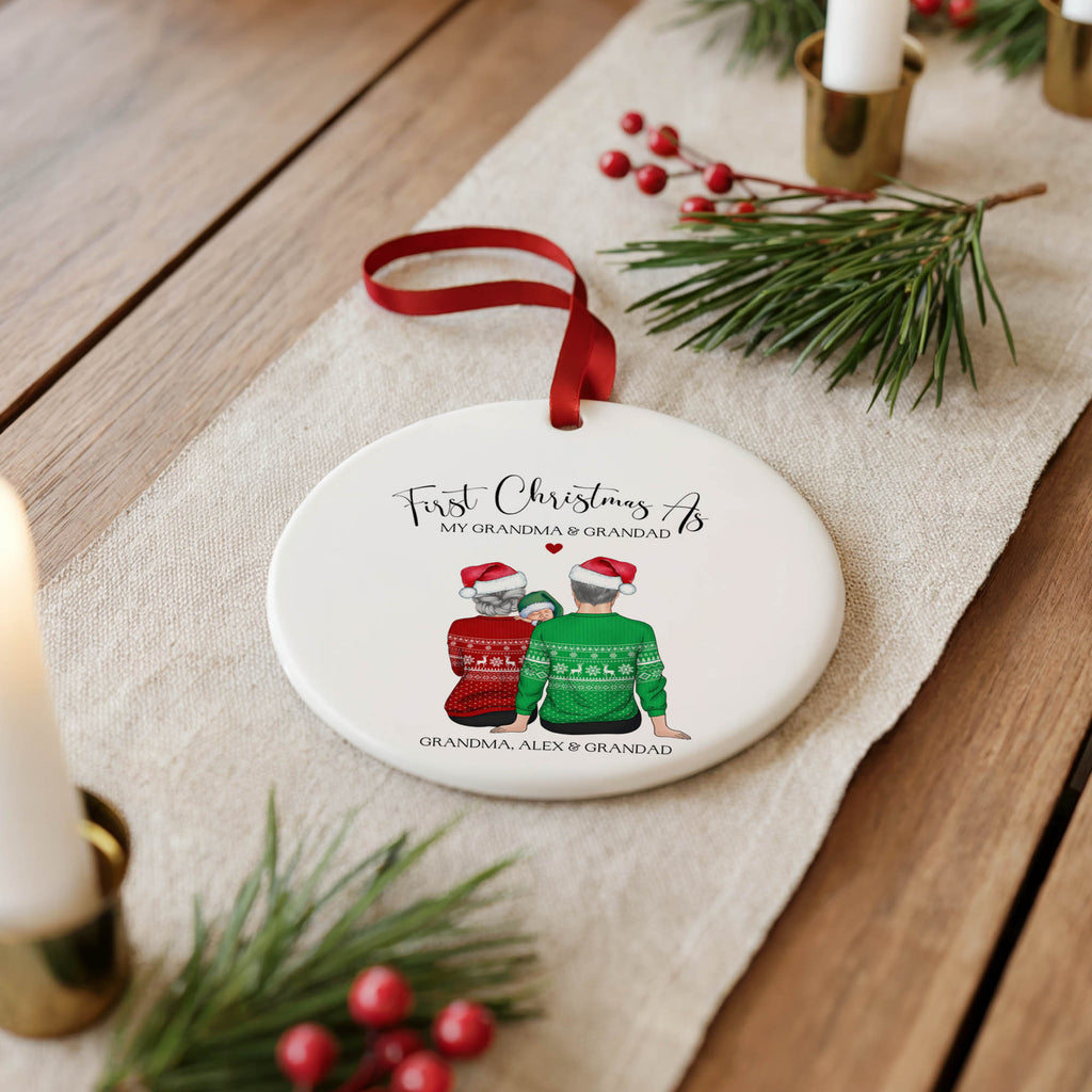 A personalized round Christmas ornament with a red ribbon hanger, featuring an illustration of two people in holiday sweaters and custom text, displayed on a wooden table with festive greenery and candles.