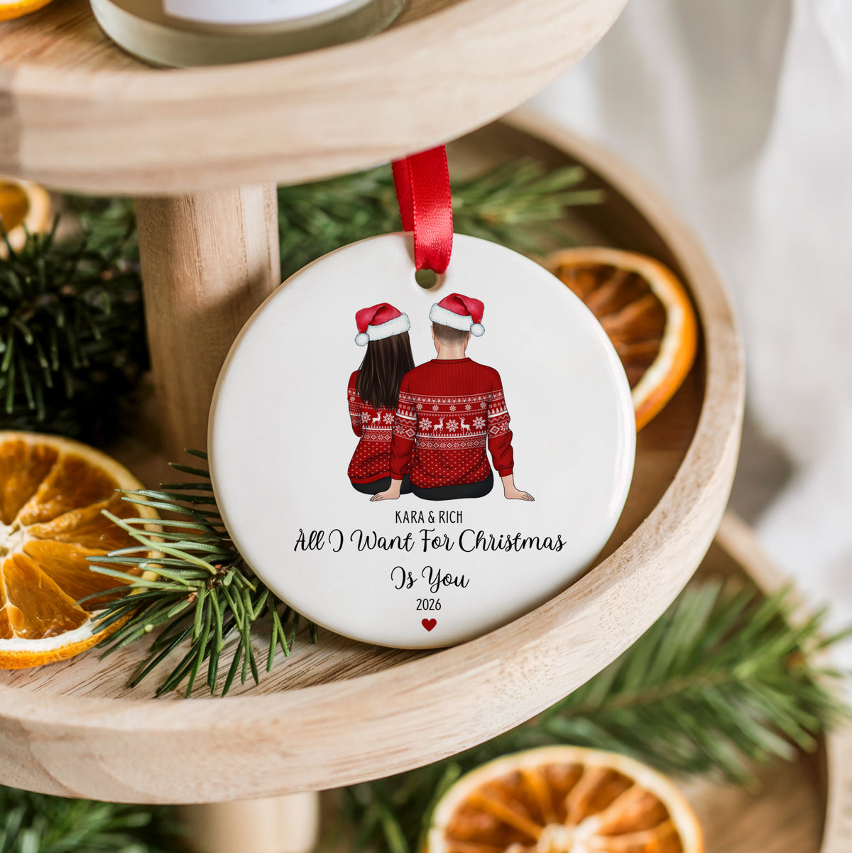 A round white ceramic Christmas ornament hanging from a red ribbon, printed with an illustration of a couple in red Santa sweaters and the personalized text 'KARA & RICH All I Want For Christmas Is You 2026'. The ornament is styled on a wooden tiered stand with pine branches and dried orange slices.
