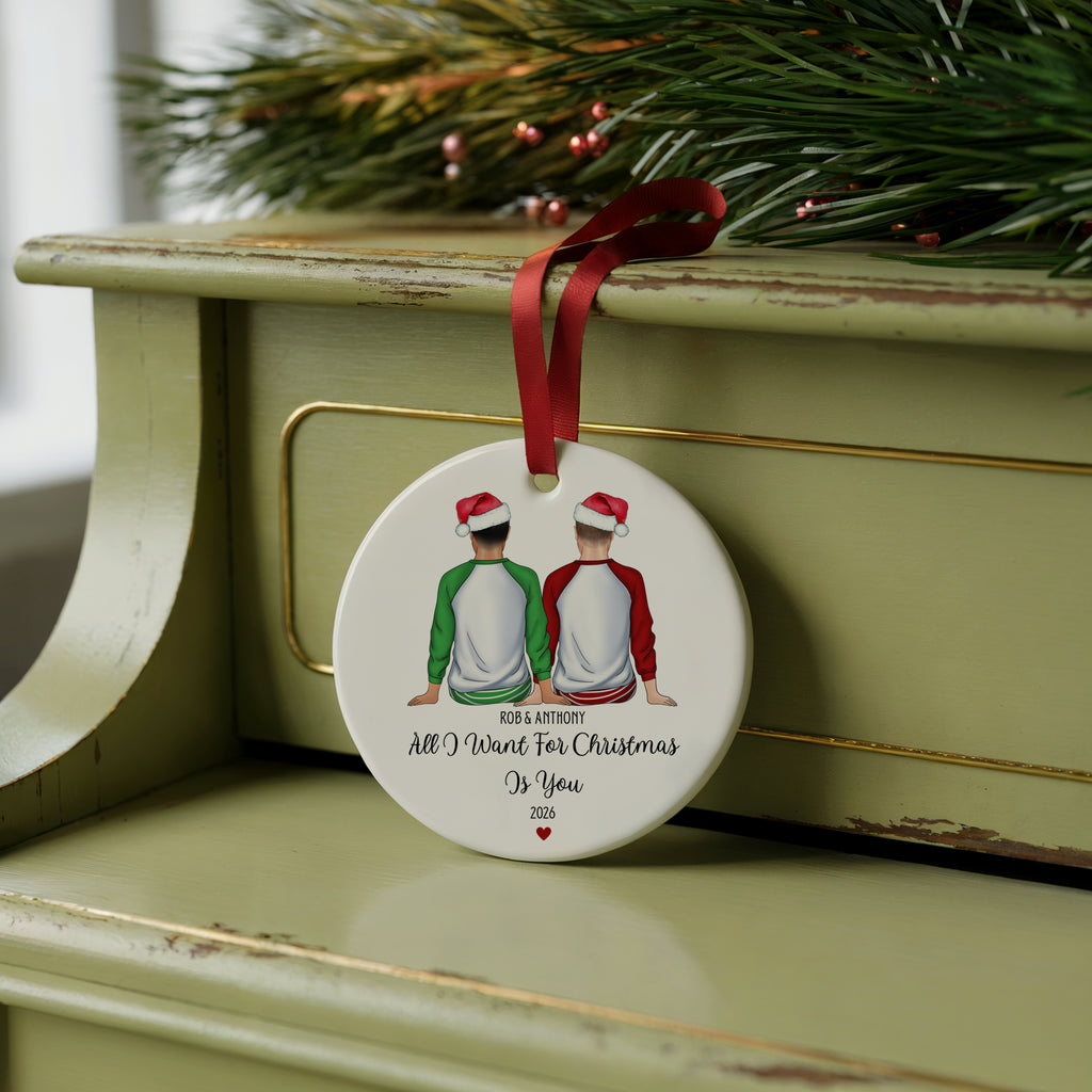 A round white Christmas ornament with a red ribbon hanging from it, featuring an illustration of two people in Santa hats sitting side by side and the printed message 'TO MY BROTHER ALL I WANT FOR Christmas Is You'. The ornament is displayed on a distressed sage green wooden surface with pine branches in the background.