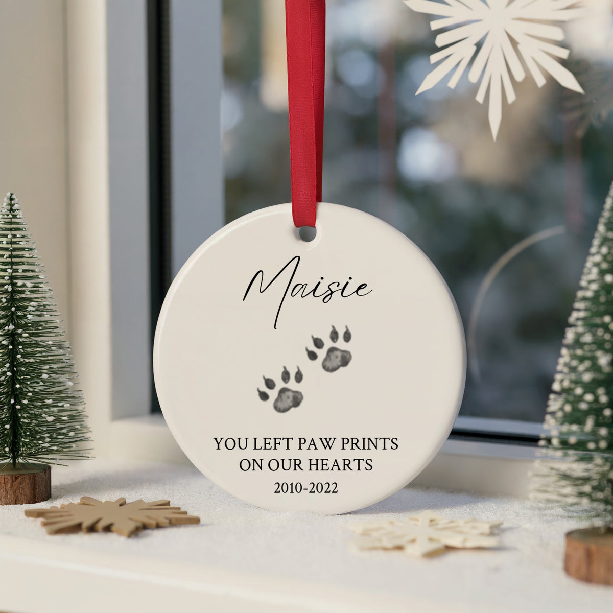 A personalized round white pet memorial ornament hanging on a window sill, featuring a red ribbon, paw prints, and the text 'Maisie YOU LEFT PAW PRINTS ON OUR HEARTS 2010-2022'. The ornament is styled with miniature trees and wooden snowflakes.