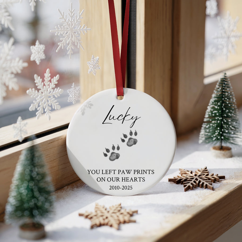 A round white ceramic Christmas ornament hanging on a wooden windowsill, printed with the name 'Lucky', paw prints, and the message 'YOU LEFT PAW PRINTS ON OUR HEARTS 2010-2025'. The ornament has a red ribbon and is surrounded by holiday decorations including small trees, wooden snowflakes, and white snowflake decals.