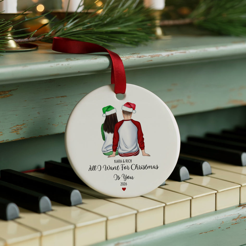 A round white Christmas ornament with a red ribbon, hanging on a distressed teal piano. The ornament features an illustration of a couple seen from behind wearing Santa hats, with the personalized text 'KABLE RICQ All I Want For Christmas Is You 2026' printed below.