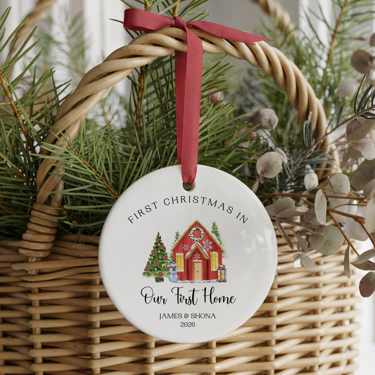 A personalized round white ceramic Christmas ornament hanging from a red ribbon inside a wicker basket with pine branches and dried foliage. The ornament is printed with the text 'FIRST CHRISTMAS IN Our First Home JAMES & SHONA 2026' above a small illustrated house, tree, and gifts.