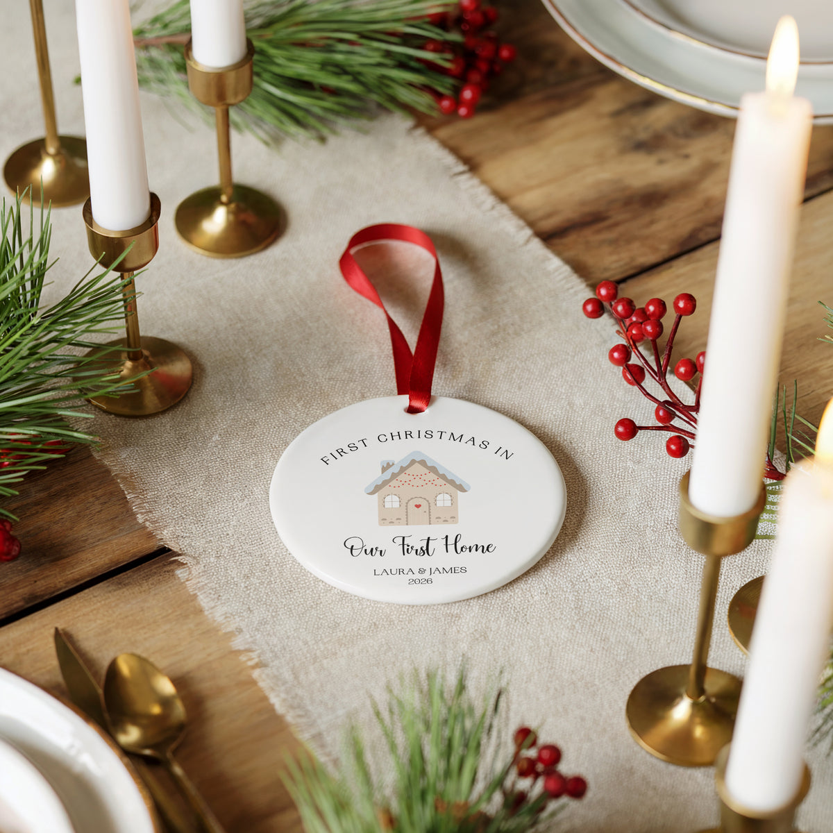 A personalized round ceramic Christmas ornament hanging on a rustic wooden table. The white ornament features a red ribbon loop and a printed illustration of a cozy house with the text 'FIRST CHRISTMAS IN Our First Home' and the name 'LAURA PLUMBER 2028'.