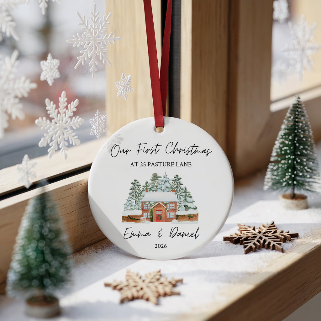 A round white Christmas ornament with a red hanging ribbon, featuring a printed illustration of a snow-covered cabin and personalized text reading 'Our First Christmas at 25 Pasture Lane Emma & Daniel 2026.' The ornament is displayed on a wooden windowsill with holiday decorations, including small trees, wooden snowflakes, and white snowflake decals.
