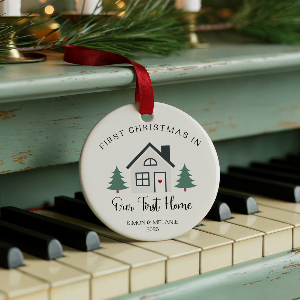 A personalized round Christmas ornament hanging from a red ribbon on a piano keyboard, with pine branches in the background. The ornament reads 'FIRST CHRISTMAS IN Our First Home SIMON & MELANIE 2023' with a small house and tree illustration.