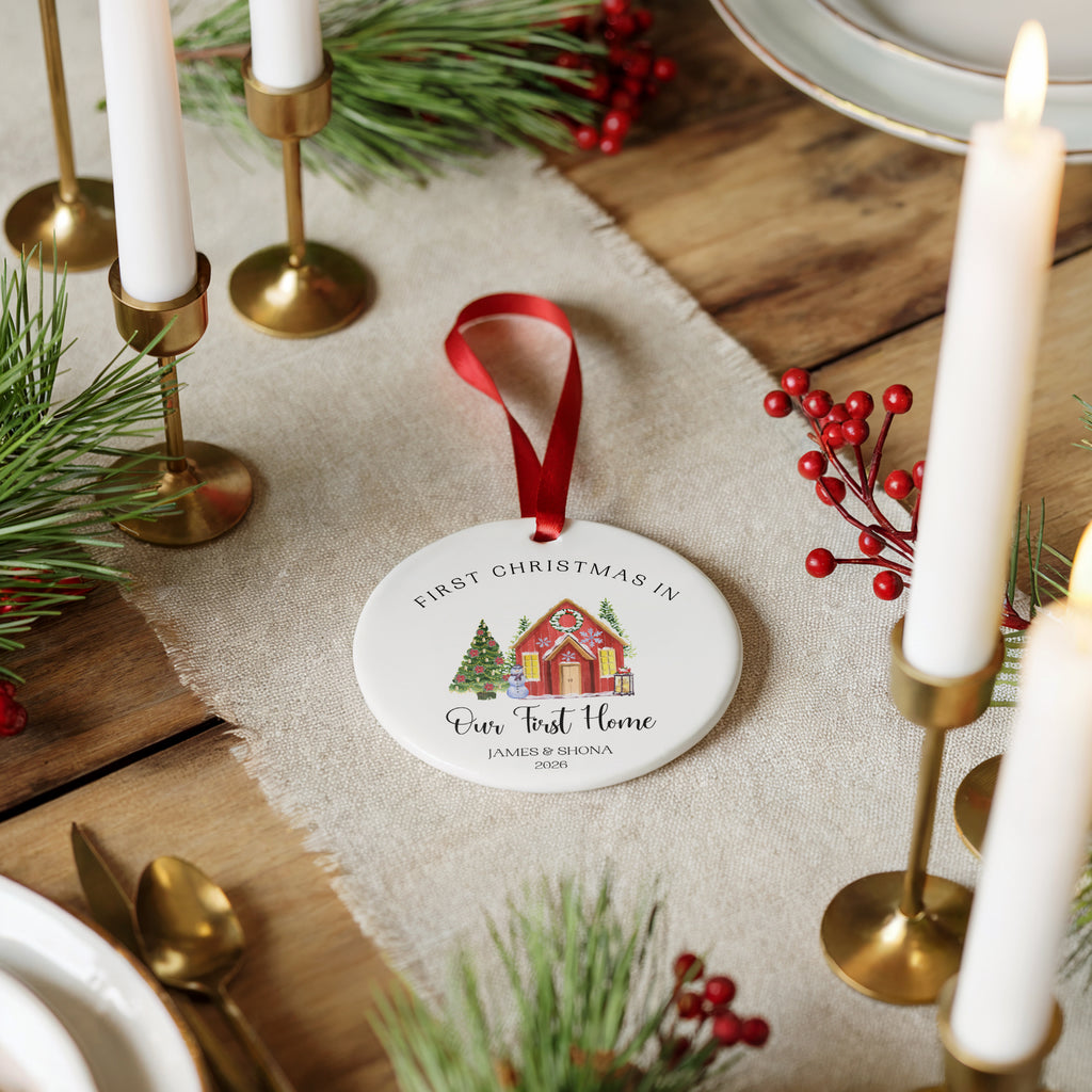 A round white ceramic Christmas ornament with a red ribbon loop, featuring a printed illustration of a holiday cottage and the text 'FIRST CHRISTMAS IN Ever Trust Home', styled on a rustic wooden table with candles, pine branches, and red berries.