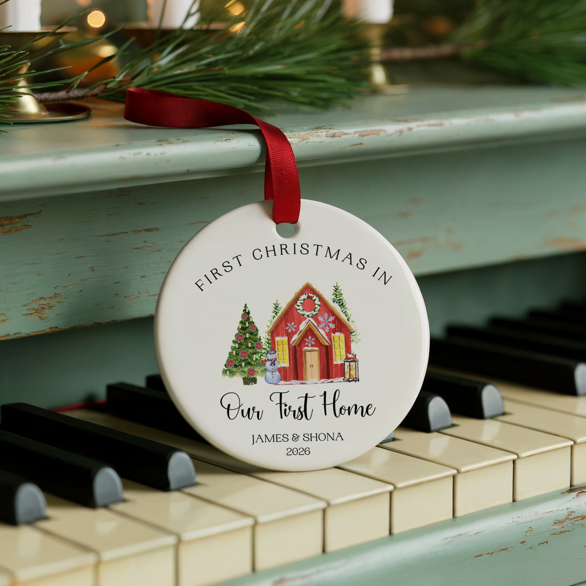A personalized round ceramic Christmas ornament hanging from a red ribbon, displayed on a vintage teal piano. The ornament features a printed illustration of a gingerbread house and a decorated Christmas tree, with the text 'FIRST CHRISTMAS IN Our First Home' and the names 'JAMES & SHONA 2026'.