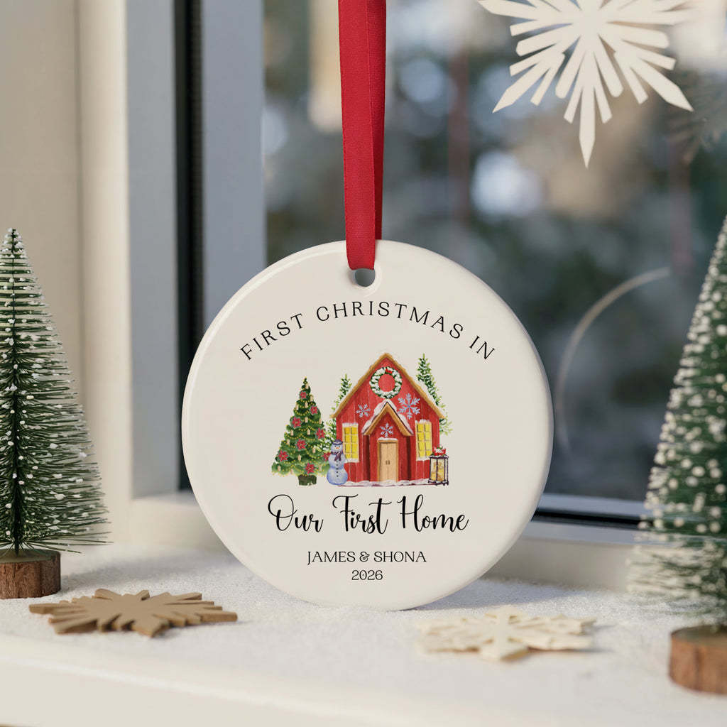 A personalized round ceramic Christmas ornament hanging on a windowsill. The white ornament features a red ribbon, the text 'FIRST CHRISTMAS IN Our First Home' and the names 'JAMES & SHONA 2026' around an illustration of a red house with a green Christmas tree.