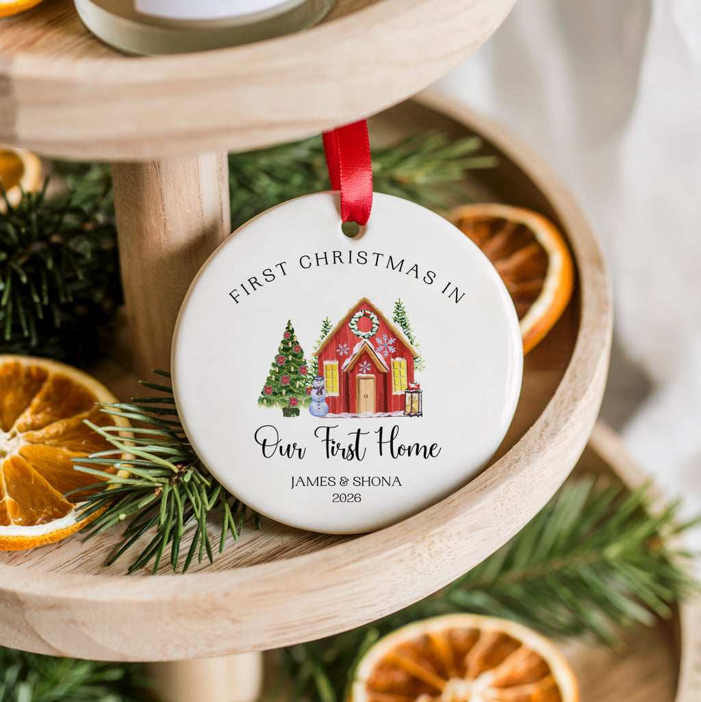 A round white personalized Christmas ornament hanging from a red ribbon, displayed on a wooden tiered stand with pine branches and dried orange slices. The ornament features a red house illustration and the text 'FIRST CHRISTMAS IN Our First Home JAMES & SHONA 2026'.