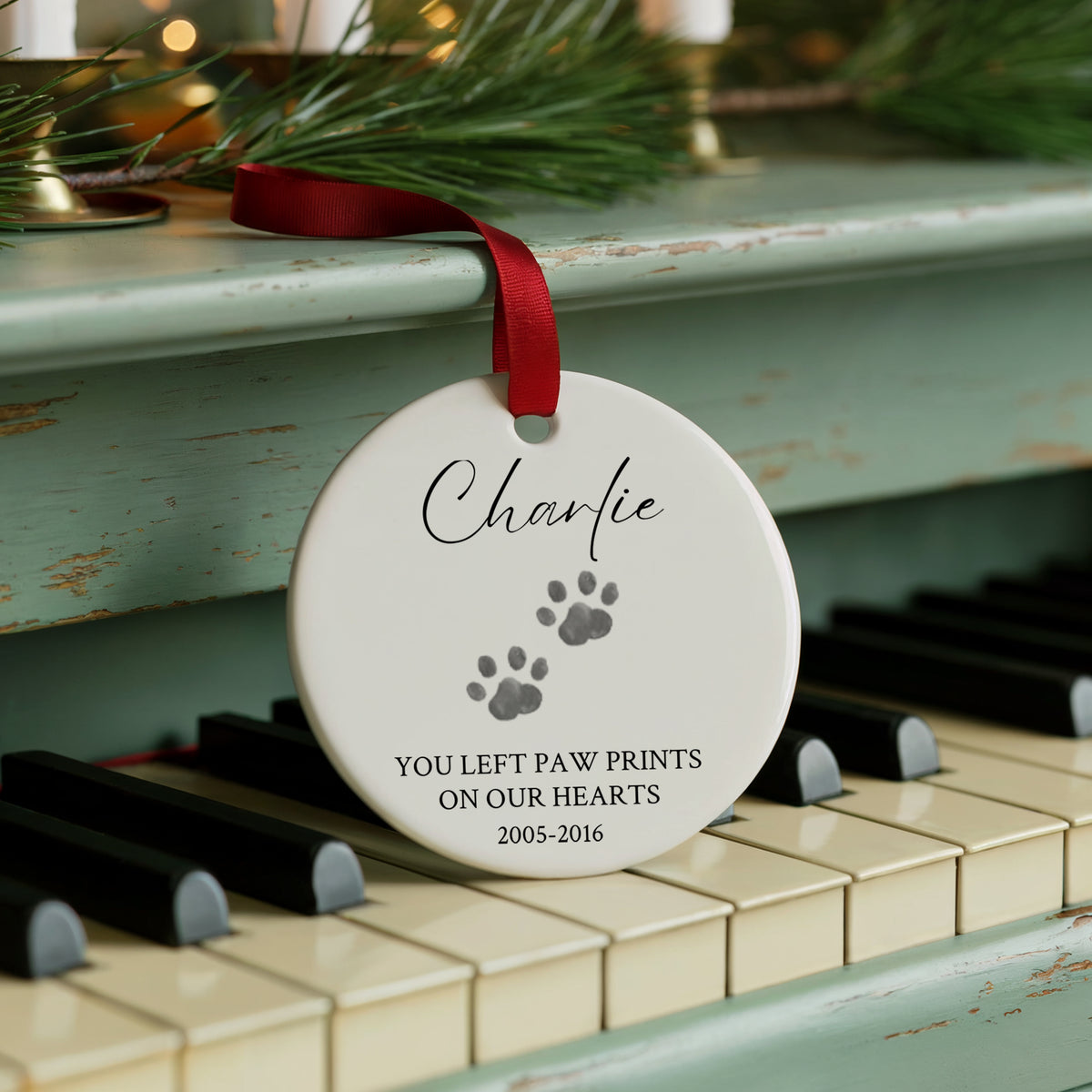 A personalized round white pet memorial ornament hanging from a red ribbon on a distressed teal piano. The ornament is engraved with the name 'Charlie', gray paw prints, and the text 'YOU LEFT PAW PRINTS ON OUR HEARTS 2005-2016'.