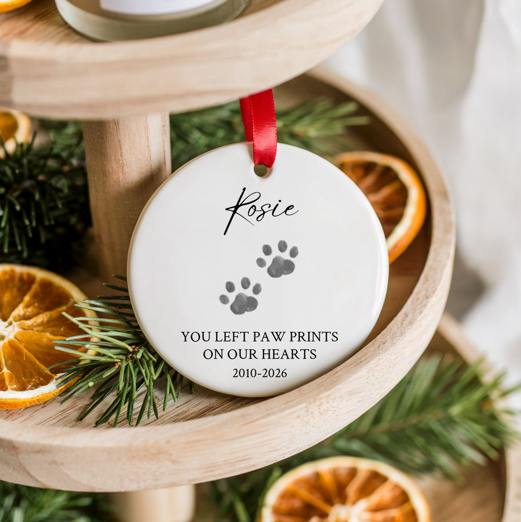A personalized round ceramic pet memorial ornament hanging on a wooden tiered stand. The white ornament is printed with the name 'Kosie', two gray paw prints, and the text 'YOU LEFT PAW PRINTS ON OUR HEARTS 2010-2026', hanging from a red ribbon.