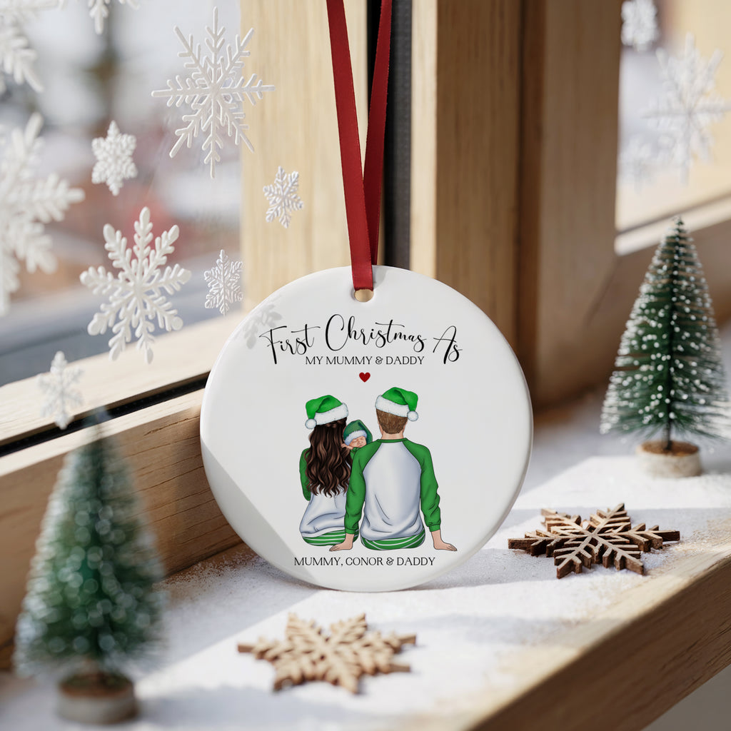 A round white Christmas ornament with a red hanging ribbon, featuring an illustration of a couple from behind wearing Santa hats, with the text 'First Christmas As MY MUMMY & DADDY' and 'MUMMY, GONNOR & DADDY'. The ornament is displayed on a wooden windowsill decorated with miniature Christmas trees and wooden snowflakes.