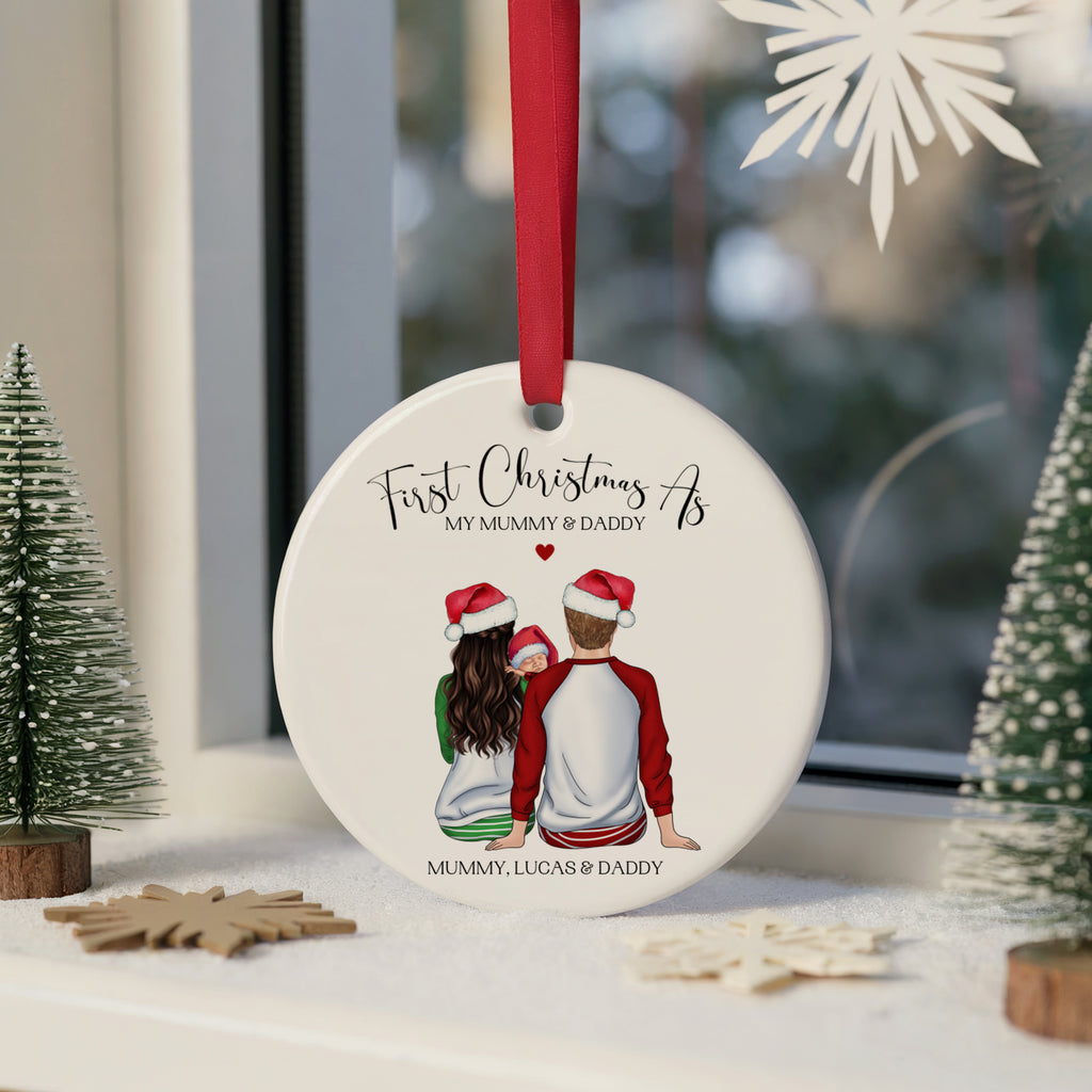 A round white ceramic Christmas ornament with a red hanging ribbon, featuring an illustration of a couple from behind wearing Santa hats, with the text 'First Christmas As MY MUMMY & DADDY' and 'MUMMY, LUCAS & DADDY'. The ornament is displayed on a windowsill with small decorative Christmas trees and wooden star ornaments.