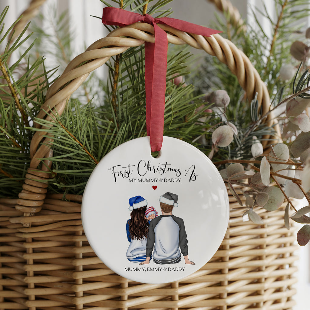 A personalized round white Christmas ornament hanging from a red ribbon inside a wicker basket with pine branches. The ornament features a watercolor illustration of a mother and father sitting with their children and the text 'First Christmas As MY Mummy & Daddy' and 'Mummy, Emmy & Daddy'.