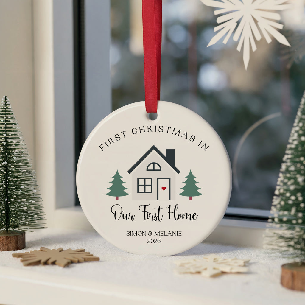 A round white ceramic Christmas ornament hanging from a red ribbon, printed with a house illustration and the text 'FIRST CHRISTMAS IN Our First Home SIMON & MELANIE 2026'. The ornament is styled on a windowsill with small green trees and wooden ornaments.