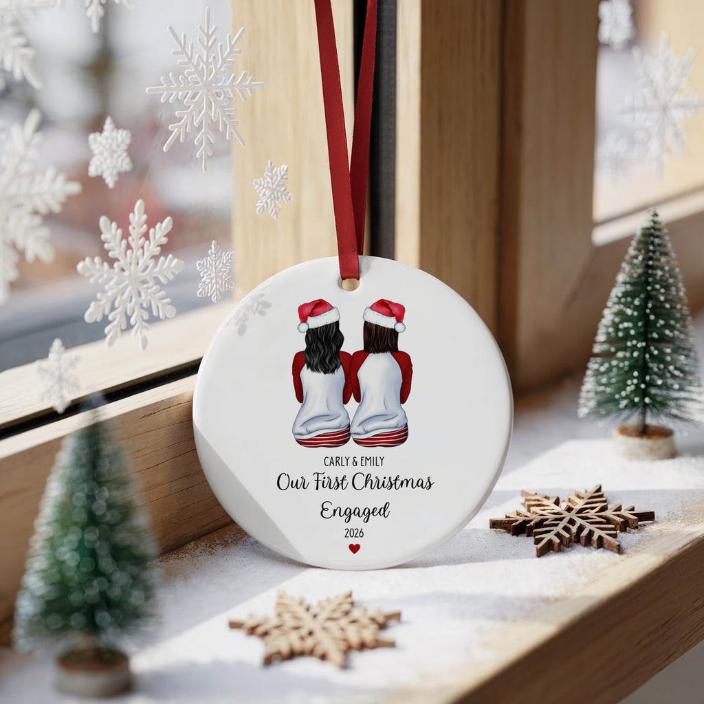 A round white Christmas ornament hanging on a wooden windowsill, featuring an illustration of two girls in Santa hats with the text 'EARY & MARY Our First Christmas Engaged 2026'. The ornament is decorated with a red ribbon and is surrounded by miniature Christmas trees, wooden snowflakes, and white snowflake decorations.