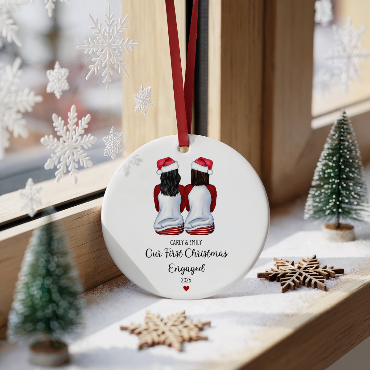 A round white Christmas ornament hanging on a wooden windowsill, featuring an illustration of two girls in Santa hats with the text 'EARY & MARY Our First Christmas Engaged 2026'. The ornament is decorated with a red ribbon and is surrounded by miniature Christmas trees, wooden snowflakes, and white snowflake decorations.