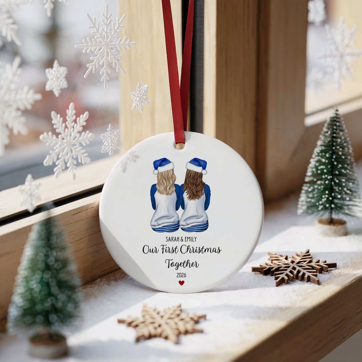 A round white ceramic Christmas ornament hanging on a wooden windowsill, featuring an illustration of two girls from behind wearing blue and white sweaters and hats, with the personalized text 'Sarah & Sammy Our First Christmas Together 2026'. The ornament is decorated with a red hanging ribbon and is surrounded by festive props including miniature trees, wooden snowflakes, and white hanging snowflakes.