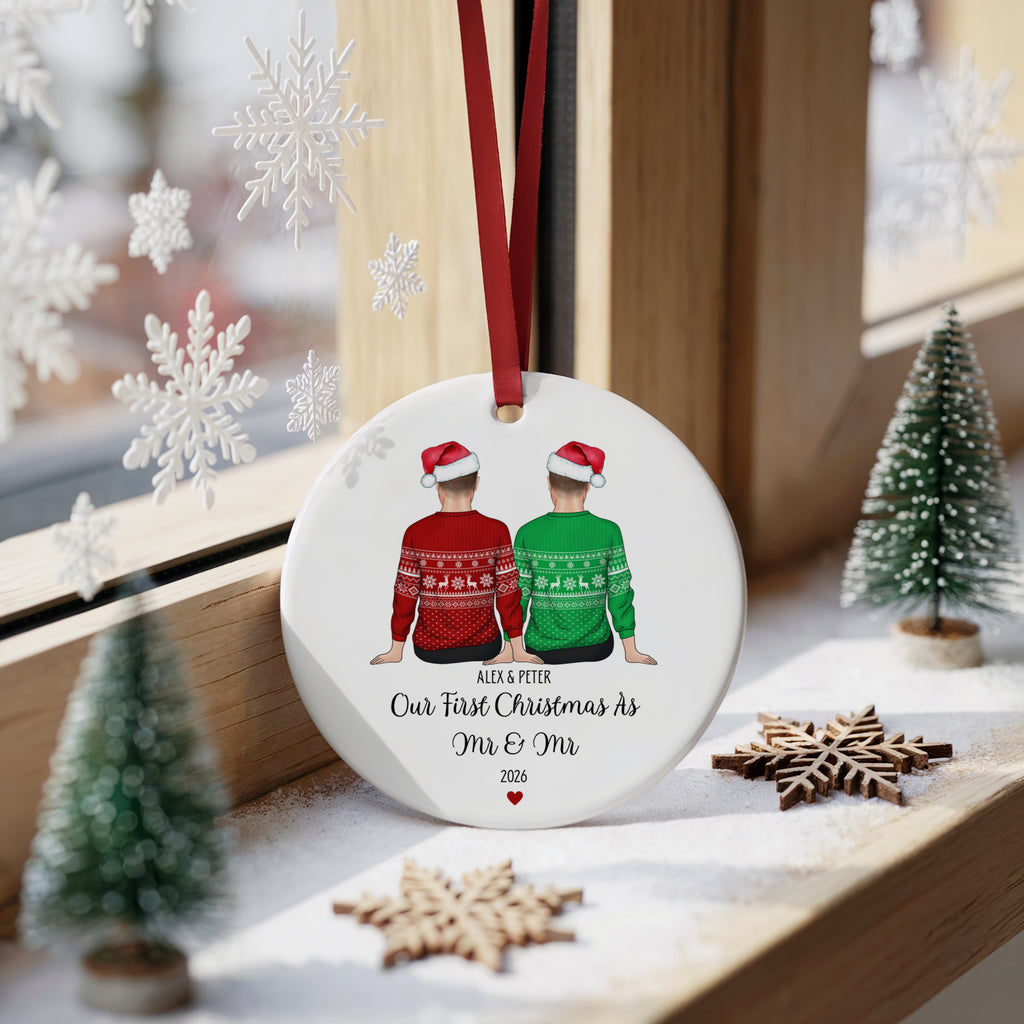 A personalized round Christmas ornament hanging on a windowsill, featuring an illustration of a couple in red and green sweaters with Santa hats. The ornament is printed with the text 'ALEX & PETER Our First Christmas As Mr & Mm 2026' and has a red hanging ribbon.