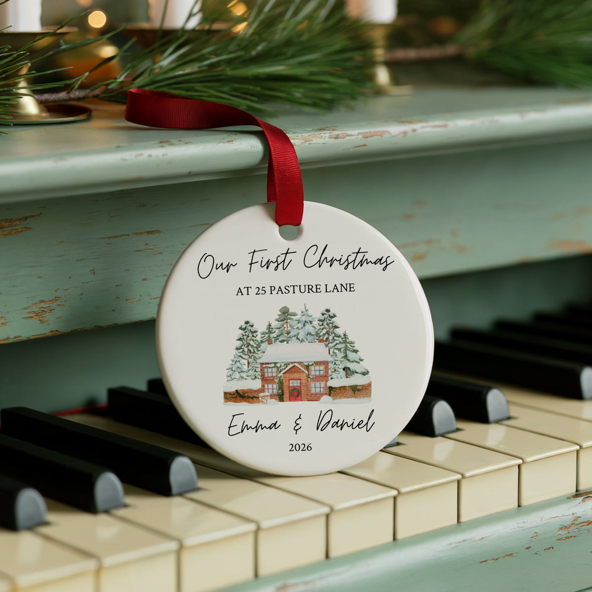 A round white ceramic Christmas ornament with a red ribbon, featuring a custom illustration and personalized text, hanging on a distressed teal piano keyboard with pine branches in the background.