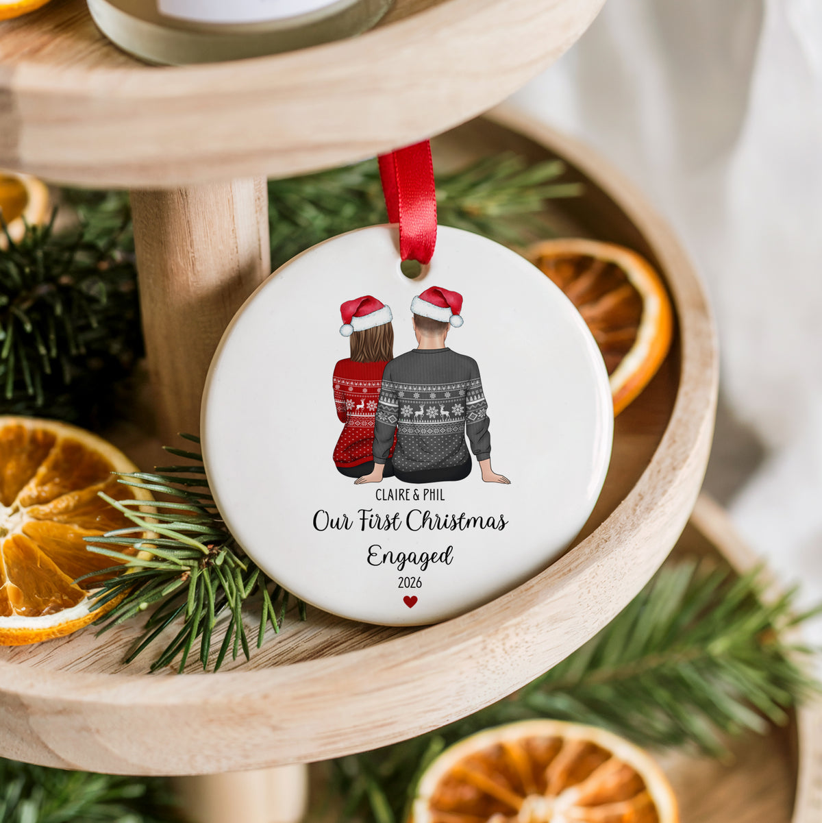 A round white ceramic Christmas ornament hanging from a red ribbon, printed with an illustration of a couple in Santa hats and the text 'Claire & Phil Our First Christmas Engaged 2026.' The ornament is styled on a wooden tiered stand with pine branches and dried orange slices.