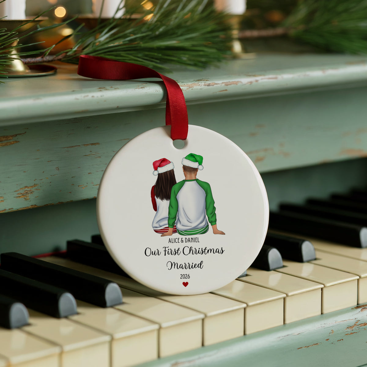 A round white Christmas ornament with a red ribbon, featuring an illustration of a couple wearing Santa hats from behind, with the text 'ALEX & RACHEL Our First Christmas Married 2026', displayed on a piano keyboard with pine branches in the background.