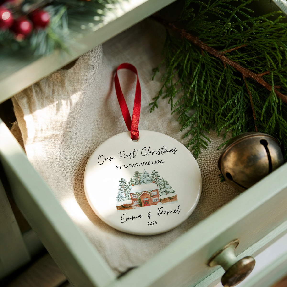 A round white ceramic Christmas ornament with a red hanging ribbon, featuring a personalized winter landscape illustration and the text 'Our First Christmas at 25 Pasture Lane Emma & Daniel 2026.' The ornament is styled inside an open drawer with pine branches, a small brass bell, and a beige fabric lining.
