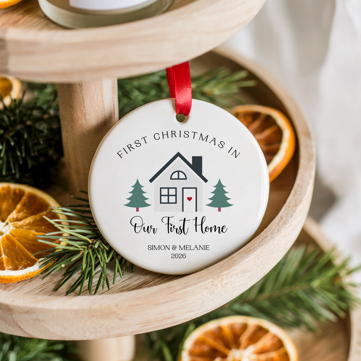 A personalized round ceramic Christmas ornament hanging from a red ribbon, displayed on a wooden tiered stand with pine branches and dried orange slices. The ornament is printed with a house illustration and the text 'FIRST CHRISTMAS IN Our First Home SIMON & MELANIE 2026'.