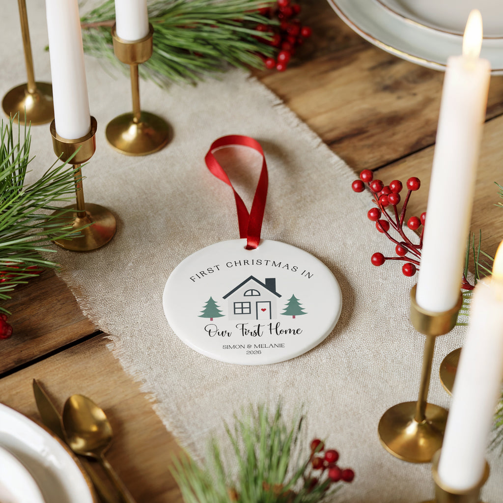 A round white ceramic Christmas ornament with a red hanging ribbon, printed with a house illustration and the text 'FIRST CHRISTMAS IN Our Trust Home,' styled on a burlap runner with candles, pine branches, and red berries on a wooden table.