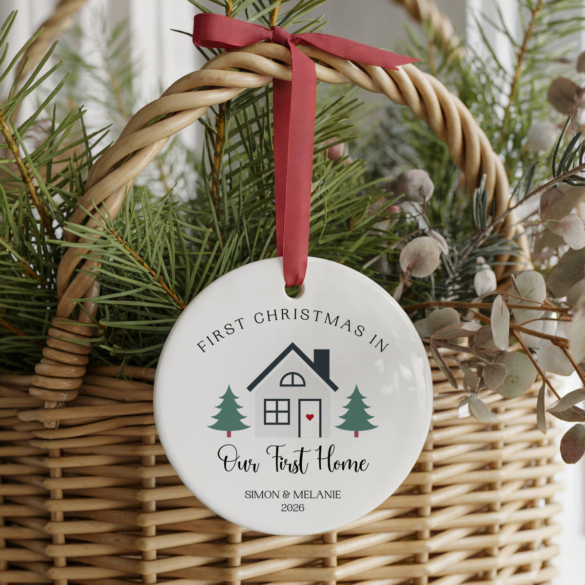 A round white ceramic Christmas ornament hanging from a red ribbon in a woven wicker basket with evergreen branches. The ornament is personalized with the text 'FIRST CHRISTMAS IN Our First Home' and the names 'SIMON & MELANIE 2026' with a small house and tree illustration.