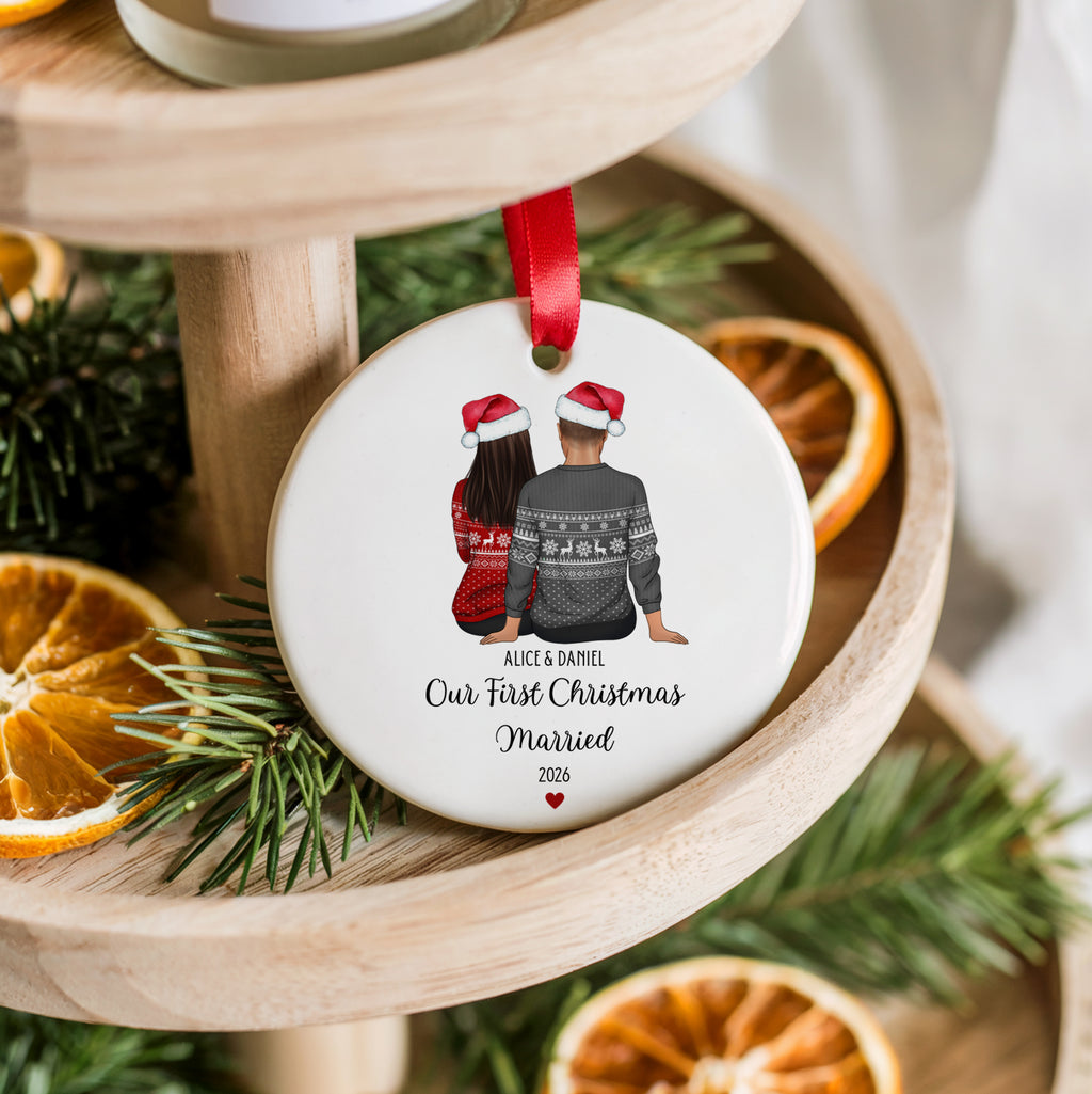 A round white Christmas ornament hanging from a red ribbon, printed with an illustration of a couple wearing Santa hats and sweaters, with the text 'Alice & Daniel Our First Christmas Married 2026'. The ornament is styled on a wooden tiered stand with pine branches and dried orange slices.