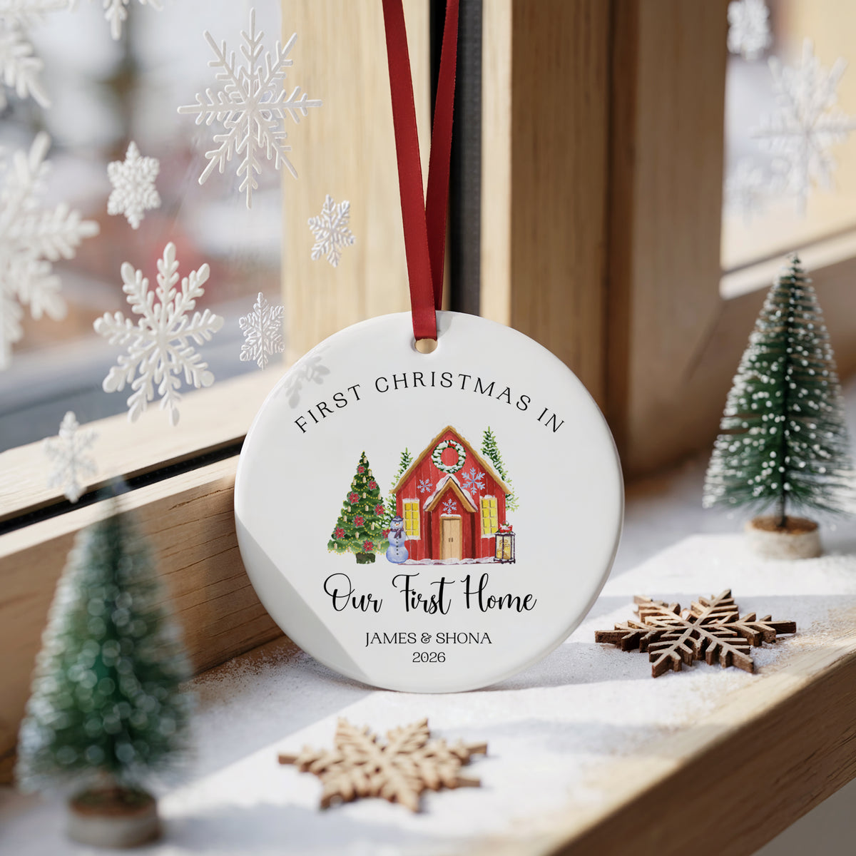 A personalized round Christmas ornament hanging on a wooden windowsill, featuring a red hanging ribbon, the text 'FIRST CHRISTMAS IN Our First Home JAMES & SHONA 2026', and a small illustrated house and Christmas trees. The scene is styled with miniature trees, wooden snowflakes, and window decorations.