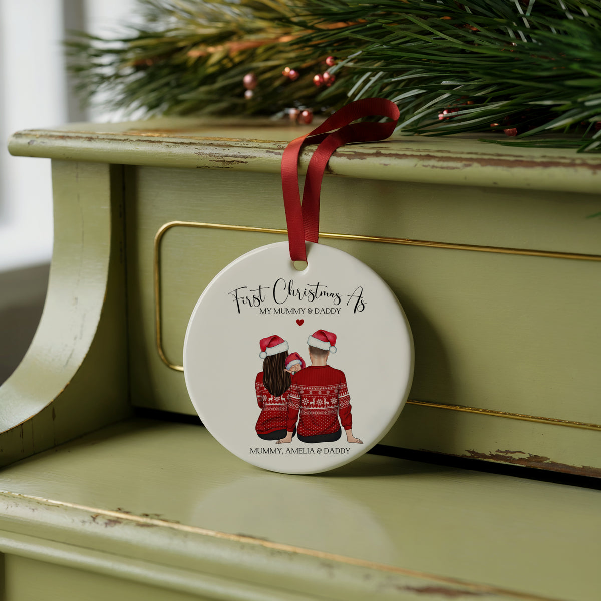 A round white Christmas ornament with a red ribbon, featuring an illustration of a mother and father in red sweaters wearing Santa hats, with the text 'First Christmas As My Legacy & Daddy' and 'MERRY, AMELIA & DADDY'. The ornament is displayed on a distressed sage green wooden chest with pine branches in the background.
