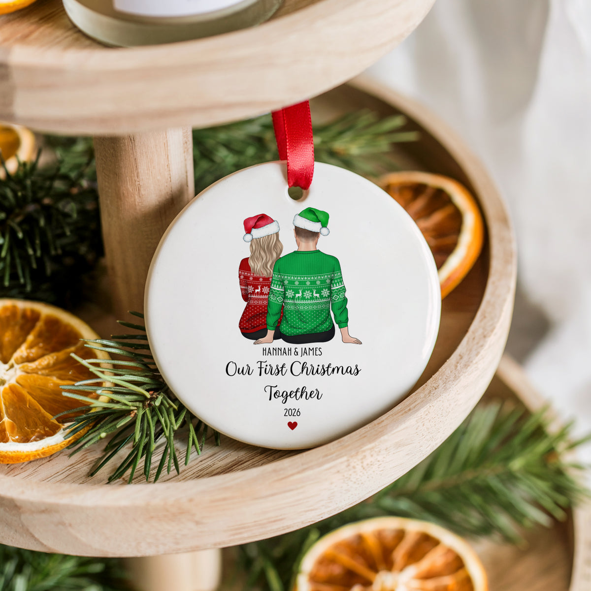 A round white ceramic Christmas ornament hanging from a red ribbon, printed with a couple in festive sweaters and the text 'HARRIS & JAMIES Our First Christmas Together 2026'. The ornament is styled on a wooden tiered stand with pine branches and dried orange slices.