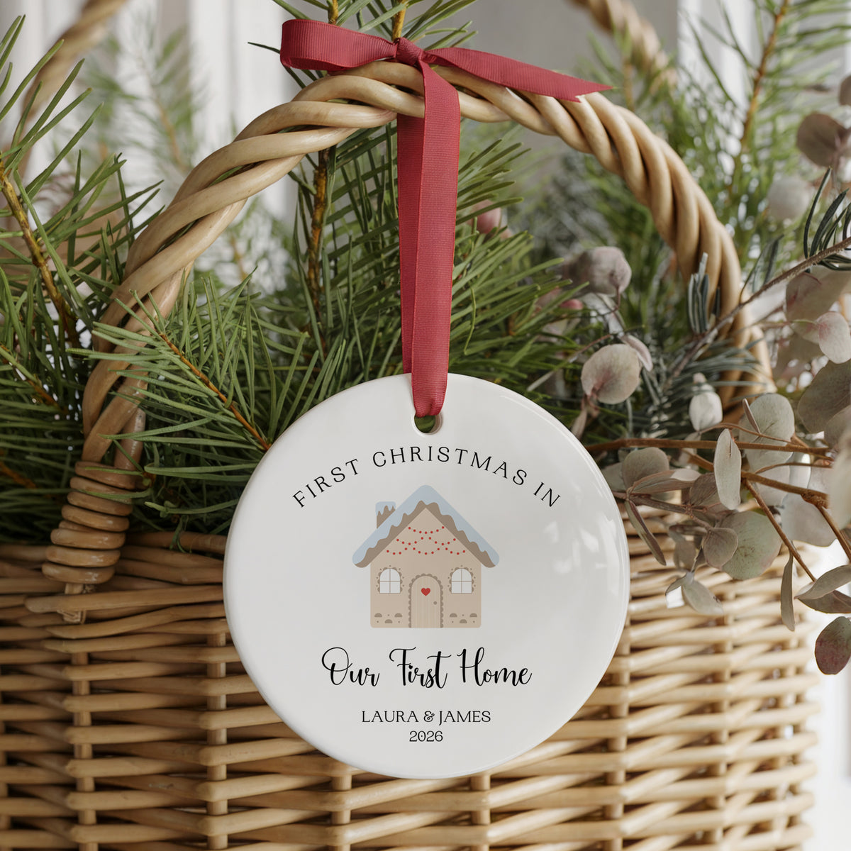 A round white ceramic Christmas ornament hanging from a red ribbon in a wicker basket with pine branches and dried foliage. The ornament is personalized with the text 'FIRST CHRISTMAS IN Our First Home LAURA & JAMES 2026' and features a small gingerbread house illustration.